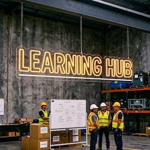 Puede incluir: Un letrero de neón rectangular que dice "LEARNING HUB" en amarillo dorado. El letrero está suspendido del techo. La imagen también muestra a personas con chalecos de seguridad y cascos, y una pizarra.