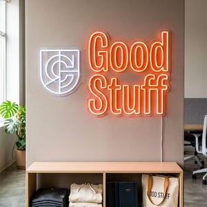 May include: A neon sign that reads "Good Stuff" in coral, with a white logo to the left. Below, a wooden shelf displays folded clothing, notebooks, and a canvas tote bag. The tote bag has the words "Good Stuff" printed on it. The sign is 36 inches wide.