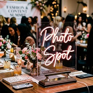 May include: A soft pink neon "Photo Spot" sign with a gold base, displayed on a wooden table. The sign is 20 inches tall and is surrounded by flowers, drinks, and people in the background. The text "THE SOCIAL SPACE • LOS ANGELES • #EventName" is below the sign.
