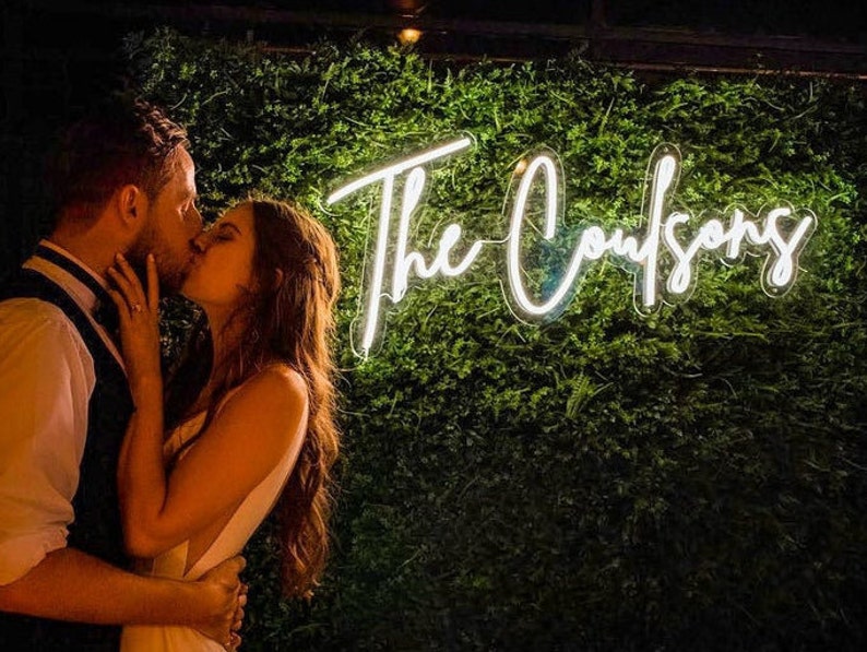 K&ouml;nnte beinhalten: Ein Paar k&uuml;sst sich vor einer gr&uuml;nen Wand mit einem wei&szlig;en Neon-Schild, auf dem "The Coufsoons" steht.
