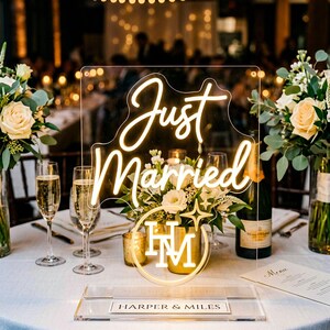 Op de afbeelding: Een helder acryl bord met warmwitte neonletters die Just Married spellen boven een cirkelvormig logo met de initialen HM en een sterpictogram. De voet bevat een rechthoekig plaatje met de gedrukte tekst Harper and Miles.