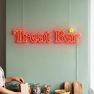 May include: A red and yellow neon sign that reads "Treat Bar" hangs above a shelf with jars of candy and brown paper bags. The sign is 36 inches wide. A person is seen opening a paper bag.