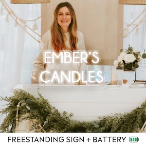Könnte beinhalten: Ein freistehendes weißes Schild mit dem Text "EMBER'S CANDLES" in neonweißem Schriftzug. Das Schild steht auf einer weißen Holztheke mit Grünpflanzen und einem batteriebetriebenen Licht.