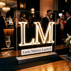 May include: A clear acrylic sign with illuminated "LM" letters and the text "Luna Maison Launch." The sign is 24 inches wide and sits on a dark wooden base. The letters glow with a warm white light, perfect for event decor.