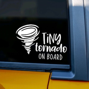 Puede incluir: Una calcomanía de vinilo blanco con un diseño de tornado estilizado y el texto "Tiny tornado on board". La calcomanía está en la ventana trasera de un coche amarillo.