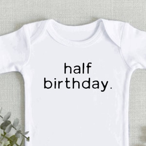 May include: White baby onesie with the text "half birthday." on the front.