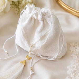 Peut inclure: Une pochette à cordon en dentelle blanche avec une fermeture froncée et des liens blancs. Un ornement est suspendu au cordon, composé d'une petite breloque papillon dorée, d'un rang de perles blanches et d'un pompon en soie blanche.