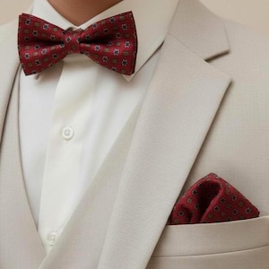 May include: A burgundy bow tie and matching pocket square featuring a repeating geometric pattern of small squares and dots. These accessories are worn with a crisp white button-down shirt and a light beige blazer with a notched lapel.