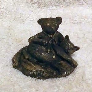 Puede incluir: Figura de color bronce de un oso de peluche sosteniendo un gato. El oso está sentado y el gato está acostado. La escultura tiene una superficie texturizada y está sobre una base texturizada.