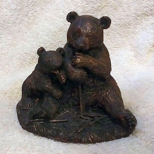 May include: A bronze-colored bear figurine depicting a mother bear and her cub. The mother bear is seated, and the cub is nestled beside her. The sculpture is detailed with textured fur and sits on a base that resembles a grassy area.