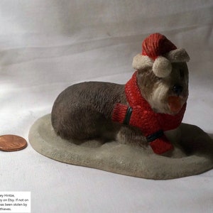 May include: A figurine of a brown and tan animal wearing a red Santa hat and scarf. The figurine is lying down on a light gray base. A penny is next to the figurine.