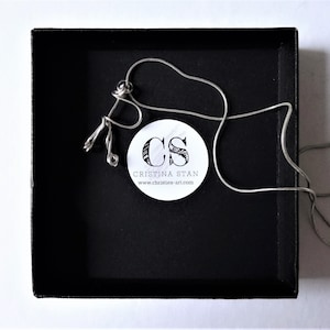 Könnte beinhalten: Eine silberfarbene Halskette mit einem einzigartigen Anhänger liegt in einer schwarzen Schachtel. Der Anhänger hat ein abstraktes Design. Ein weißer Aufkleber mit dem Text "CRISTINA STAN" und der Website-Adresse ist sichtbar.