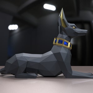 K&ouml;nnte beinhalten: Eine schwarze Anubis-Statue aus Origami mit goldenen Ohren und einem blauen Schmuckhalsband. Die Statue liegt auf einer hellgrauen Oberfl&auml;che.