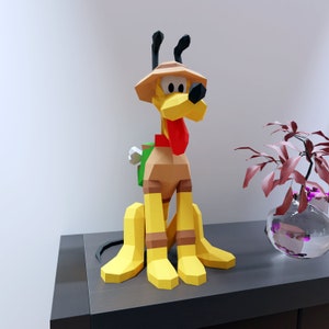 Könnte beinhalten: Ein Papiermodell von Pluto, dem Hund aus Disneys Mickey Mouse-Cartoons. Pluto trägt einen braunen Hut und einen grünen Rucksack. Er steht auf einer dunkelgrauen Oberfläche.