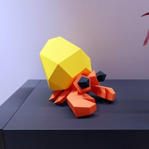 May include: A yellow and orange papercraft hermit crab with black eyes. The crab is sitting on a dark gray surface.