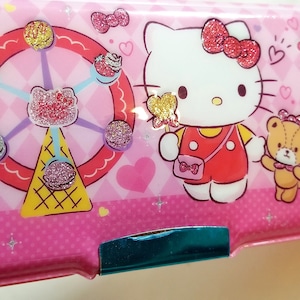 May include: Pink Hello Kitty pencil case with a Ferris wheel, a bear, and a gift box design. The case has a zipper closure and a turquoise trim.