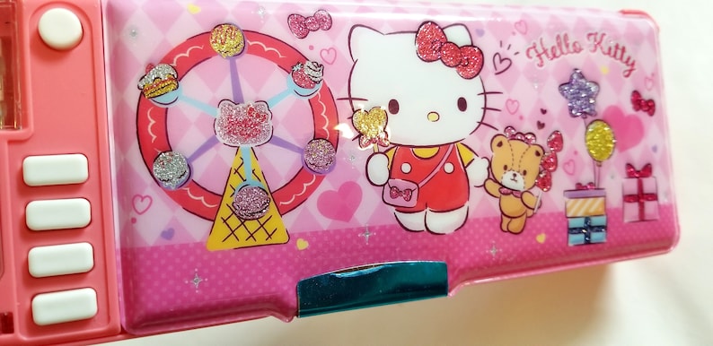 May include: Pink Hello Kitty pencil case with a Ferris wheel, a bear, and a gift box design. The case has a zipper closure and a turquoise trim.