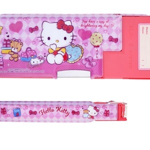 May include: Pink Hello Kitty pencil case with a clear plastic front and a pink and white checkered pattern. The case has a sliding compartment and a built-in pencil sharpener. The case features a Hello Kitty design with a pink bow and a heart. The text "You have a way of brightening my day!" is printed on the case. The text "Multy 5 system" is printed on the side of the case.