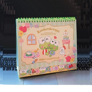 May include: A 2026 calendar featuring Kero Kero Keroppi characters. The calendar has a green spiral binding and a light green background with floral and character illustrations. The calendar includes the text "Kerokerokeroppi" and "2026 CALENDAR".