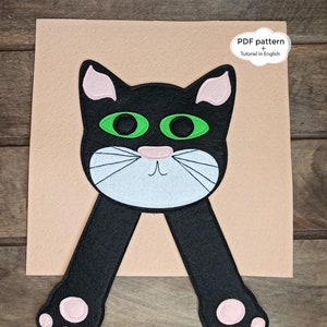 May include: A black felt cat with pink ears and paws, green eyes, and a white face. The cat is hanging from the top of the image with its arms outstretched. The background is a light brown felt.