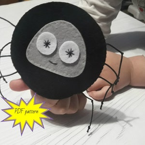 Puede incluir: Una araña de fieltro negro con una cara gris y ojos blancos. La araña tiene ocho patas de cuerda negra y una boca sonriente. El texto "PDF pattern" está en una estrella amarilla.