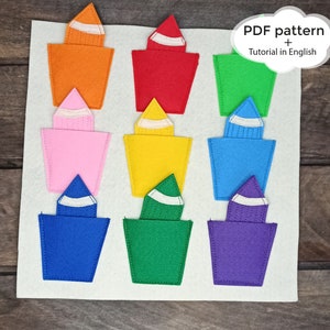Puede incluir: Un tablero de fieltro con nueve formas de crayón de fieltro en diferentes colores: naranja, rojo, rosa, amarillo, azul, verde, morado y blanco. Los crayones de fieltro están dispuestos en tres filas de tres. El tablero tiene el texto "PDF pattern + Tutorial in English" en una forma de nube blanca.
