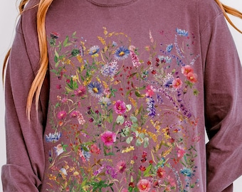 May include: Berry-colored long-sleeve shirt featuring a vibrant floral design. The graphic includes a variety of colorful flowers and foliage. The shirt has a crew neck and long sleeves. The word "berry" is printed at the bottom.