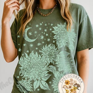 May include: Moss green t-shirt featuring a floral and celestial design. The design includes a crescent moon, stars, and large flowers in a lighter green hue. The shirt has a relaxed fit and short sleeves. The Coco Bear original design logo is visible.