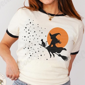 May include: Cream ringer t-shirt with black trim, featuring a Halloween design. The graphic shows a witch on a broom with a cat, stars, and an orange moon. The text "natural/black" is at the bottom right.