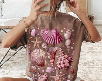 Puede incluir: Una camiseta marrón con estampado de conchas rosas y blancas. La camiseta presenta una variedad de conchas, incluyendo una gran concha de vieira, una concha de caracol y una estrella de mar.