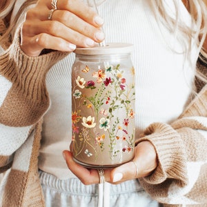 May include: A glass jar with a floral design and a straw. The jar is filled with a brown liquid and has a lid. The jar is being held by a person wearing a white tank top and a brown and white cardigan.