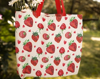 May include: A white canvas tote bag with red straps and a repeating pattern of red strawberries and pink hearts. The bag is hanging from a tree branch and has a bouquet of white daisies inside.
