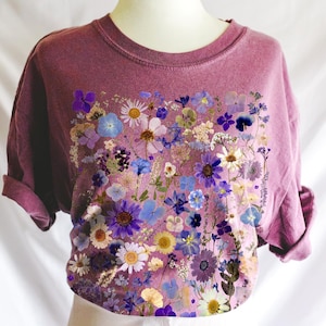 May include: A mauve t-shirt with a floral print design. The print features a variety of dried flowers in shades of purple, blue, and white.