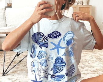 May include: A light blue t-shirt featuring a graphic print of blue and white seashells and a starfish. The shirt is made from a soft, comfortable fabric and has a relaxed fit.