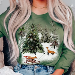May include: A green long-sleeved shirt with a winter scene featuring a Christmas tree, snow, and two foxes. One fox is pulling a sled and the other is standing in the snow.