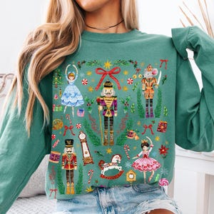 May include: Light green sweatshirt with a festive Nutcracker design. The illustration features nutcrackers, ballerinas, and a rocking horse. The text "light green" is visible in the bottom right corner. The sweatshirt has long sleeves.