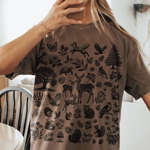 Può includere: T-shirt color espresso con un'illustrazione nera dettagliata di animali e piante del bosco. Il design include cervi, volpi, uccelli, alberi e fiori. La maglietta ha una vestibilità ampia e maniche corte.