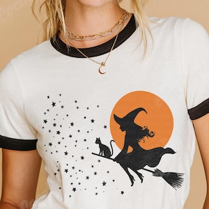 以下が含まれることがあります： 黒い縁取りの白いTシャツ。ハロウィーンのデザインが特徴です。グラフィックは、黒猫、星、オレンジ色の月と一緒にほうきに乗った魔女を描いています。カジュアルな普段着です。