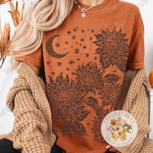 Pode incluir: Uma camiseta cor de ferrugem com um design floral e celestial. O gr&aacute;fico inclui uma lua crescente, estrelas e grandes ilustra&ccedil;&otilde;es de flores em um tom marrom mais escuro. A camiseta faz parte da cole&ccedil;&atilde;o "The Coco Bear Original Design".