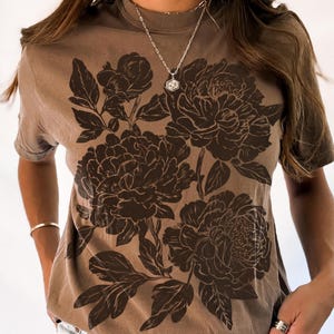 May include: Espresso-colored t-shirt featuring a large floral design in a darker brown hue. The graphic depicts detailed peony blooms and leaves. The shirt has a crew neck and short sleeves. The word "espresso" is printed at the bottom left.