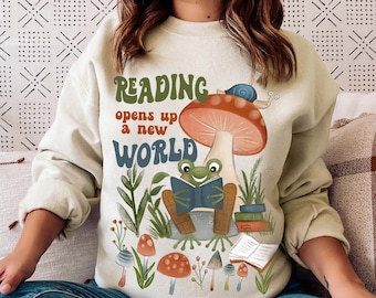 Può includere: Una felpa beige chiaro con una rana verde che legge un libro sotto un fungo rosso e bianco. Il testo "READING opens up a new WORLD" è stampato sulla felpa.