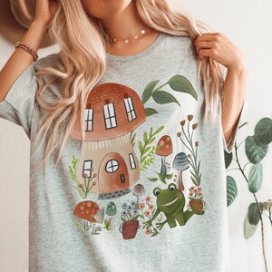 K&ouml;nnte beinhalten: Ein graues Oversize-T-Shirt mit einer fantasievollen Illustration eines Pilzhauses, eines Frosches und Pilzen in einer Gartenlandschaft.