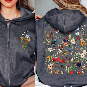 May include: A dark heather grey zip-up hoodie with a floral print on the back and a smaller floral print on the left chest. The hoodie has a drawstring hood.