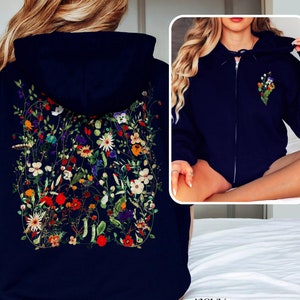 May include: Navy blue women's zip-up hoodie featuring a vibrant floral print on the back. The print showcases a variety of colorful flowers and greenery. The hoodie has a classic design with a drawstring hood and kangaroo pocket. The word 'navy' is visible at the bottom of the image.