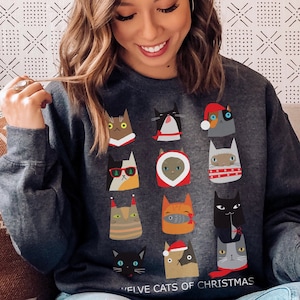 Può includere: Una felpa grigia scura con un design festivo che presenta dodici gatti vestiti con vari abiti natalizi. Il testo "Twelve Cats of Christmas" è stampato nella parte inferiore della felpa.