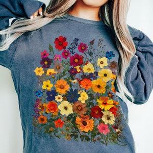Könnte beinhalten: Ein blaues Sweatshirt mit einem bunten Blumenmuster. Die Blumen sind gepresst und in Form eines Straußes angeordnet. Das Sweatshirt hat eine lockere Passform und einen Rundhalsausschnitt.