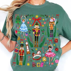 May include: Light green t-shirt with a festive Nutcracker ballet design. The illustration includes a Christmas tree, a ballerina, a nutcracker soldier, and a mouse king, surrounded by gifts and candy canes, perfect for the holiday season.