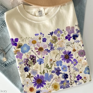 May include: A cream-colored t-shirt with a floral print featuring a variety of purple, blue, and white flowers. The shirt has the text "COMFORT COLORS" printed on the front.