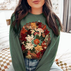 May include: A green long-sleeved sweatshirt with an embroidered autumn-themed design featuring leaves, mushrooms, and flowers.