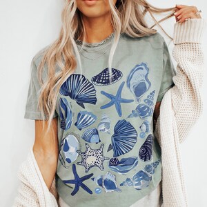 Puede incluir: Una camiseta verde claro con un estampado de conchas marinas azules y blancas. La camiseta presenta una variedad de conchas, incluyendo estrellas de mar, almejas y ostras.